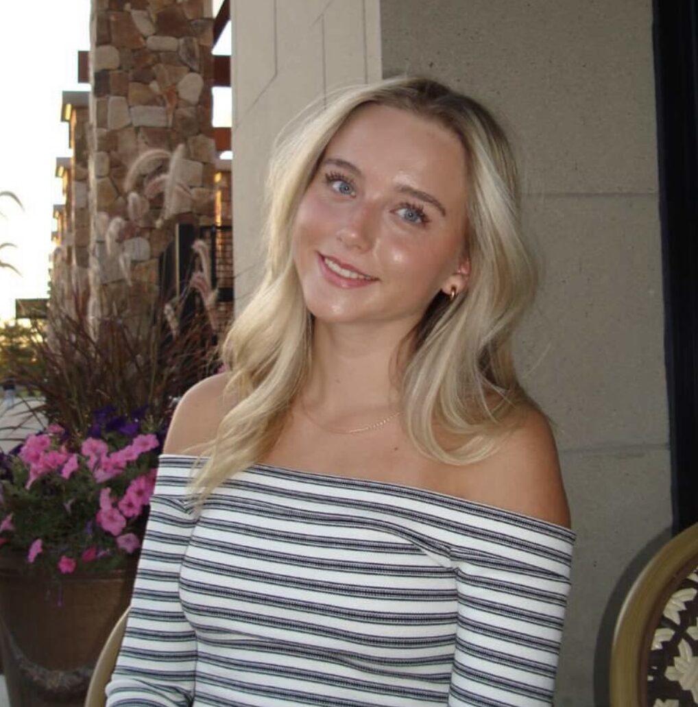 Young woman smiling in a striped off-the-shoulder top, sitting in front of a floral arrangement, representing a fresh perspective in home design and window treatments.
