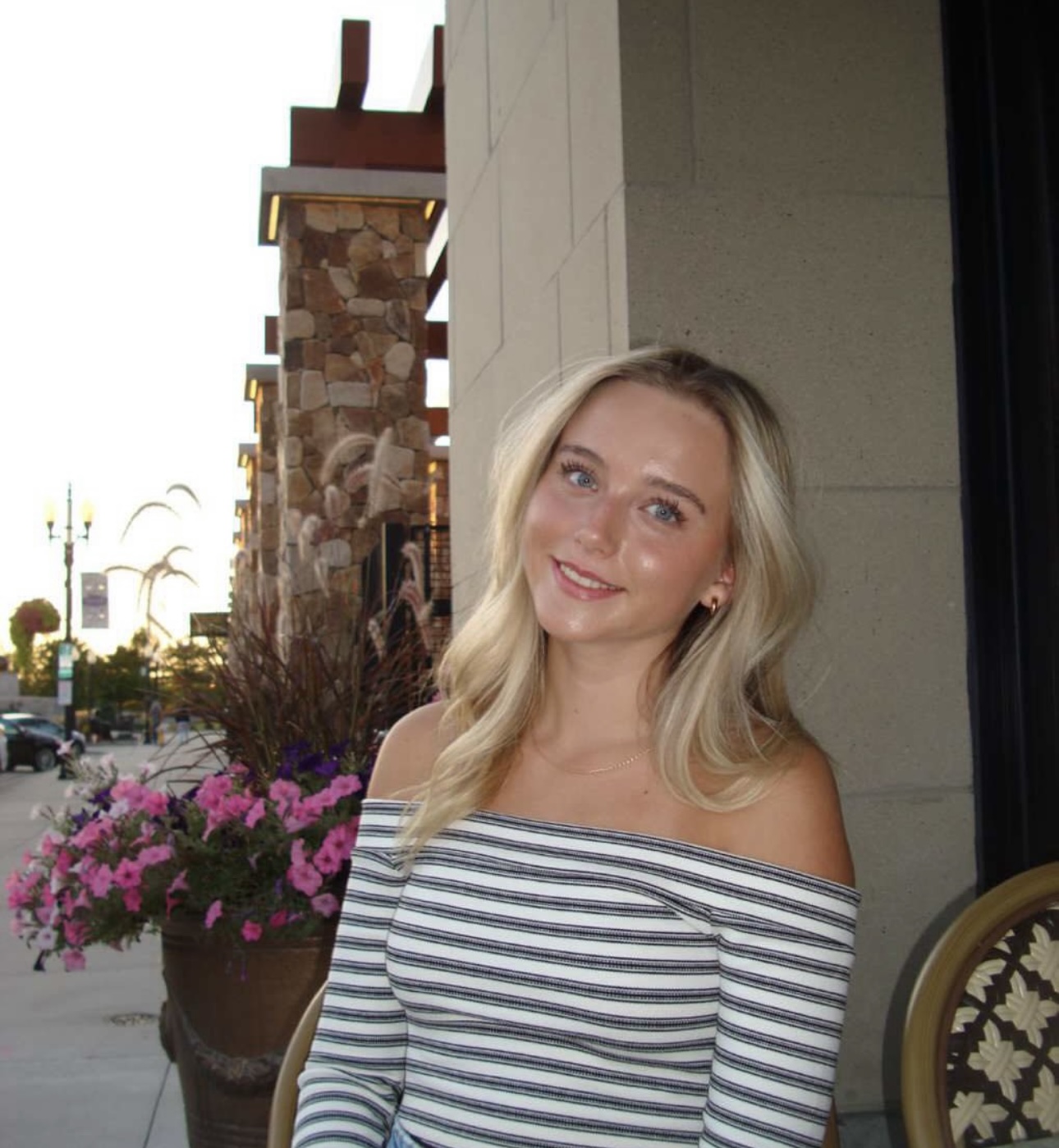 Young woman with long blonde hair, wearing a striped off-the-shoulder top, smiling while seated near a flower pot with pink flowers, in a stylish outdoor setting.