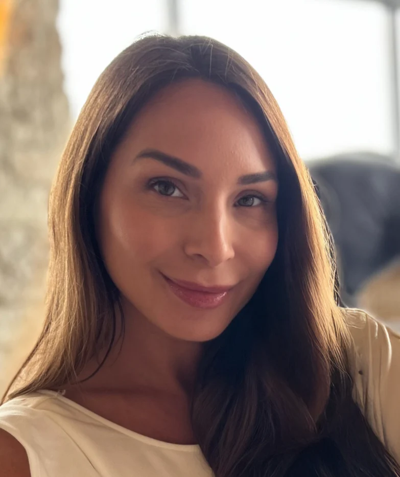 Woman with long hair smiling, wearing a white top, in a bright and inviting interior setting, representing the personalized service and design expertise of Blinds by Valeri.